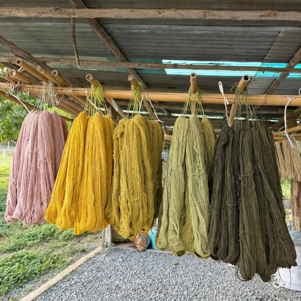 Several large bundled skeins of yarn or thread hang from bamboo rods under a rustic corrugated metal roof. The bundles are shown in muted earthy tones including dusty pink, mustard yellow, olive green, and dark brown, with grass and gravel visible around the structure.