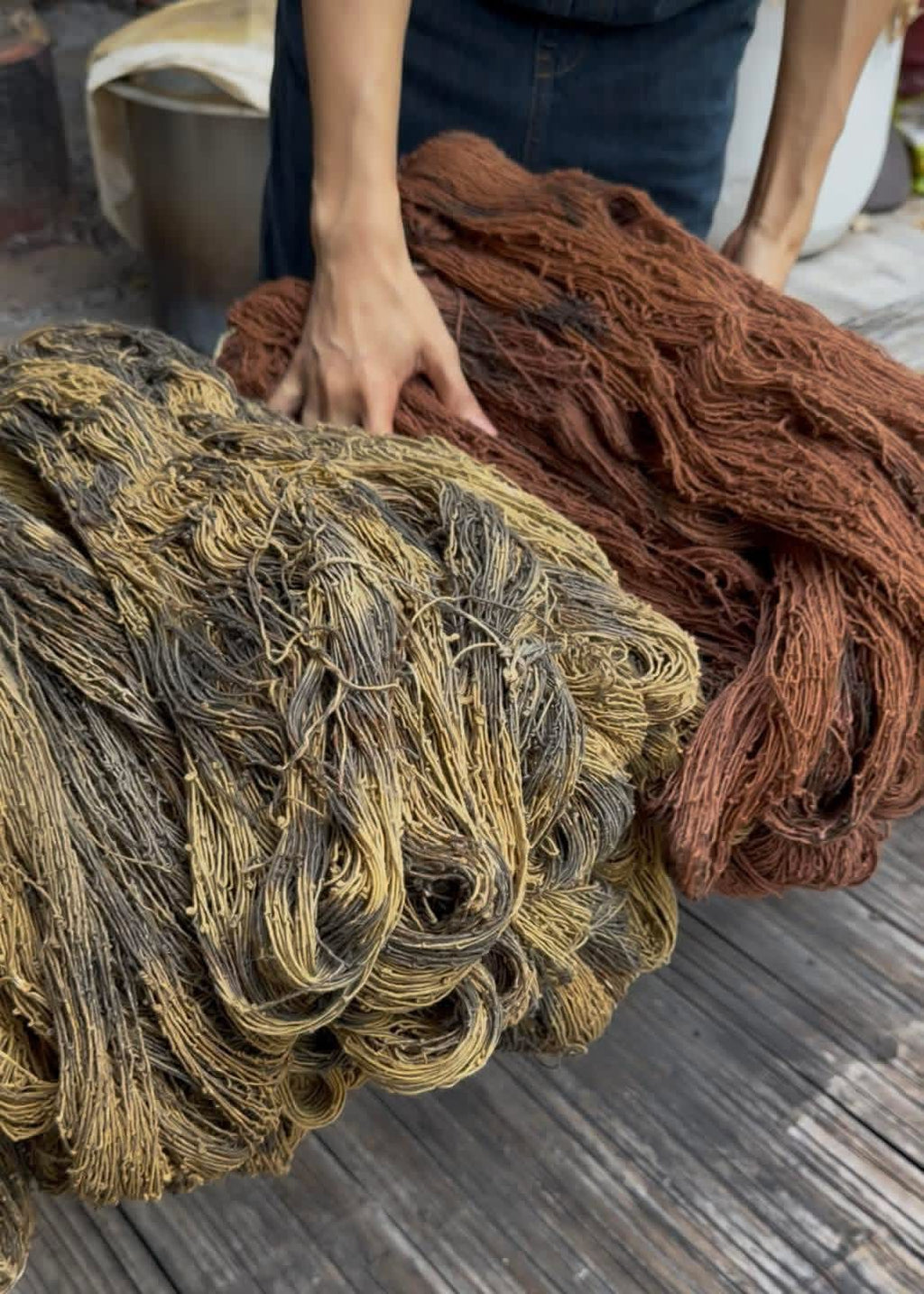 Two large bundles of thick twisted fiber yarn or rope, one in dark brown and one in tan and brown, held by a person on a weathered wooden deck.