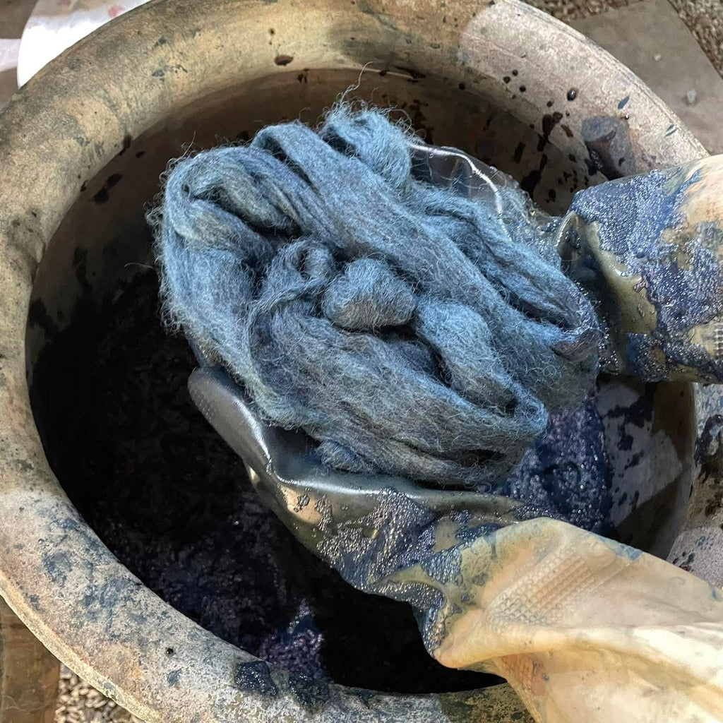 A close-up of a person's gloved hands holding a bundle of indigo fiber inside a large, worn ceramic pot filled with dark liquid.