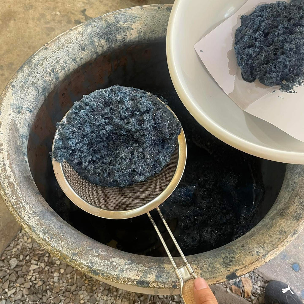 A close-up of an Indigo Forest fibrous material being lifted into a metal strainer over a large rustic vessel, with more of the same material in a white bowl nearby.