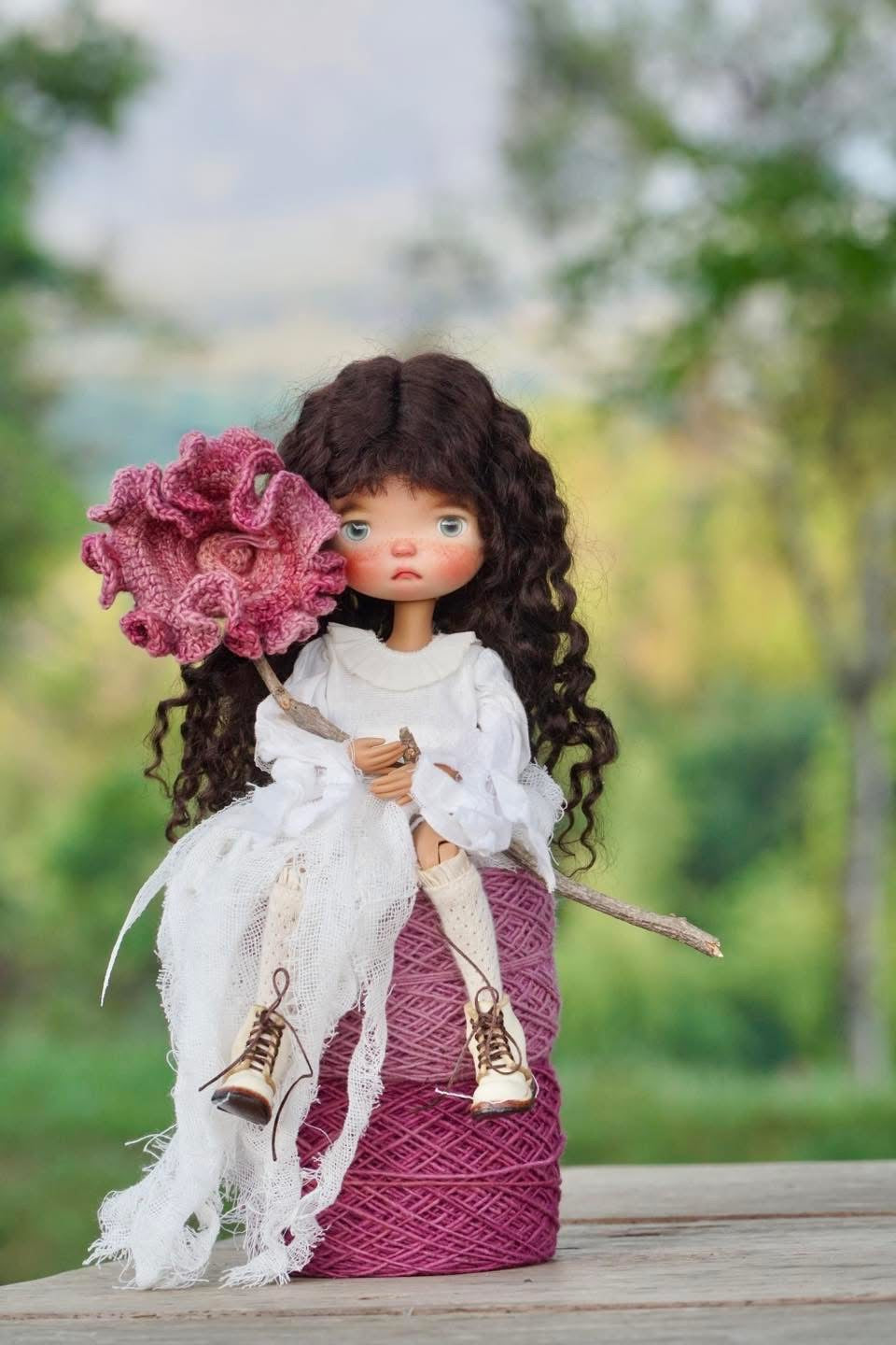 A doll with long curly brown hair sits on a wooden surface outdoors, wearing a white dress and holding a large pink flower and a small stick, with a blurred green garden background.