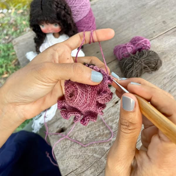 Close-up of hands crocheting with a wooden crochet hook and pink yarn, with balls of yarn and a doll in the background on a wooden surface.
